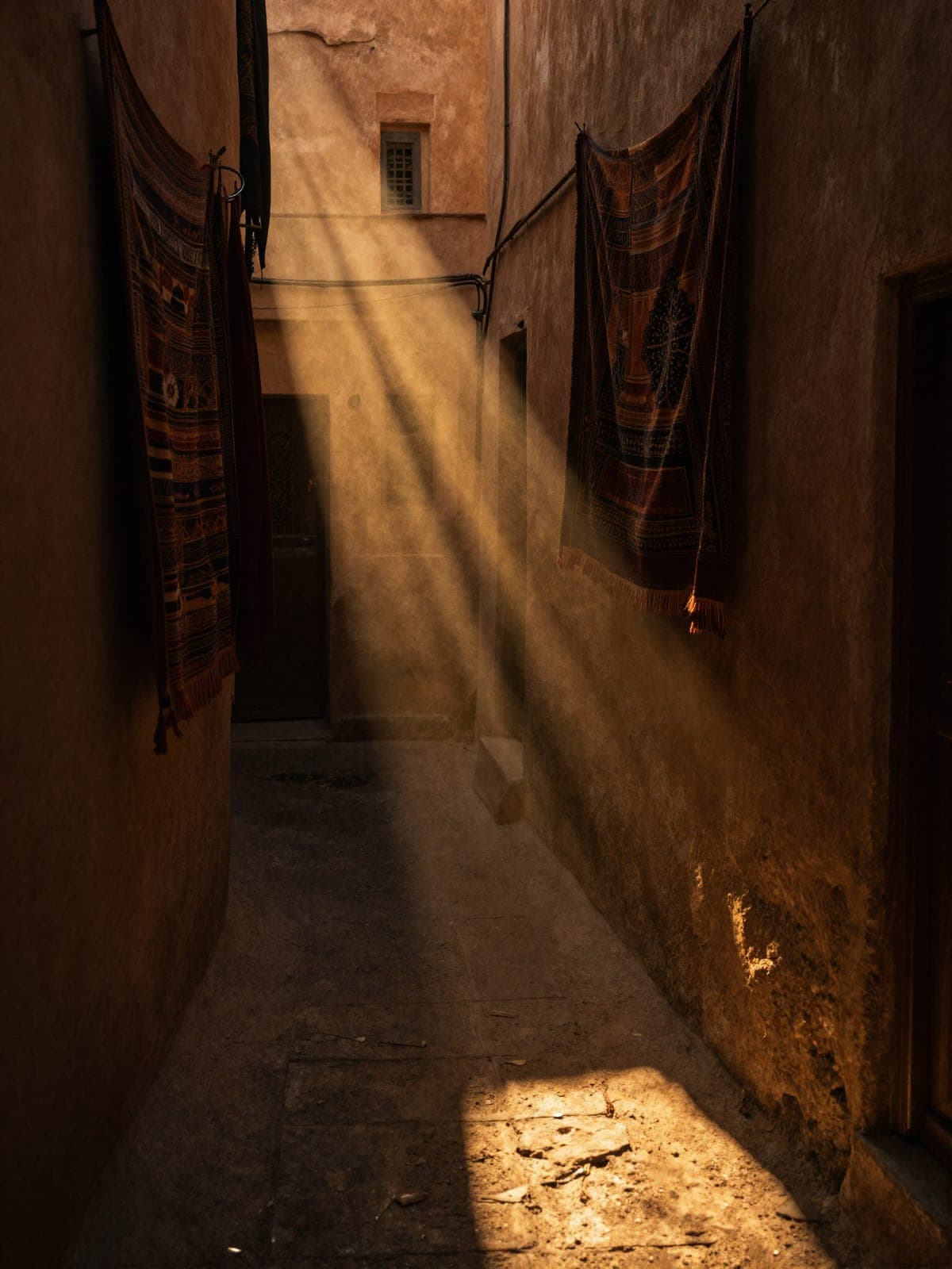 A shaft of low sun cutting down a narrow ochre alley in a medina, catching dust in the air, with two woven rugs hung out to air along the walls.