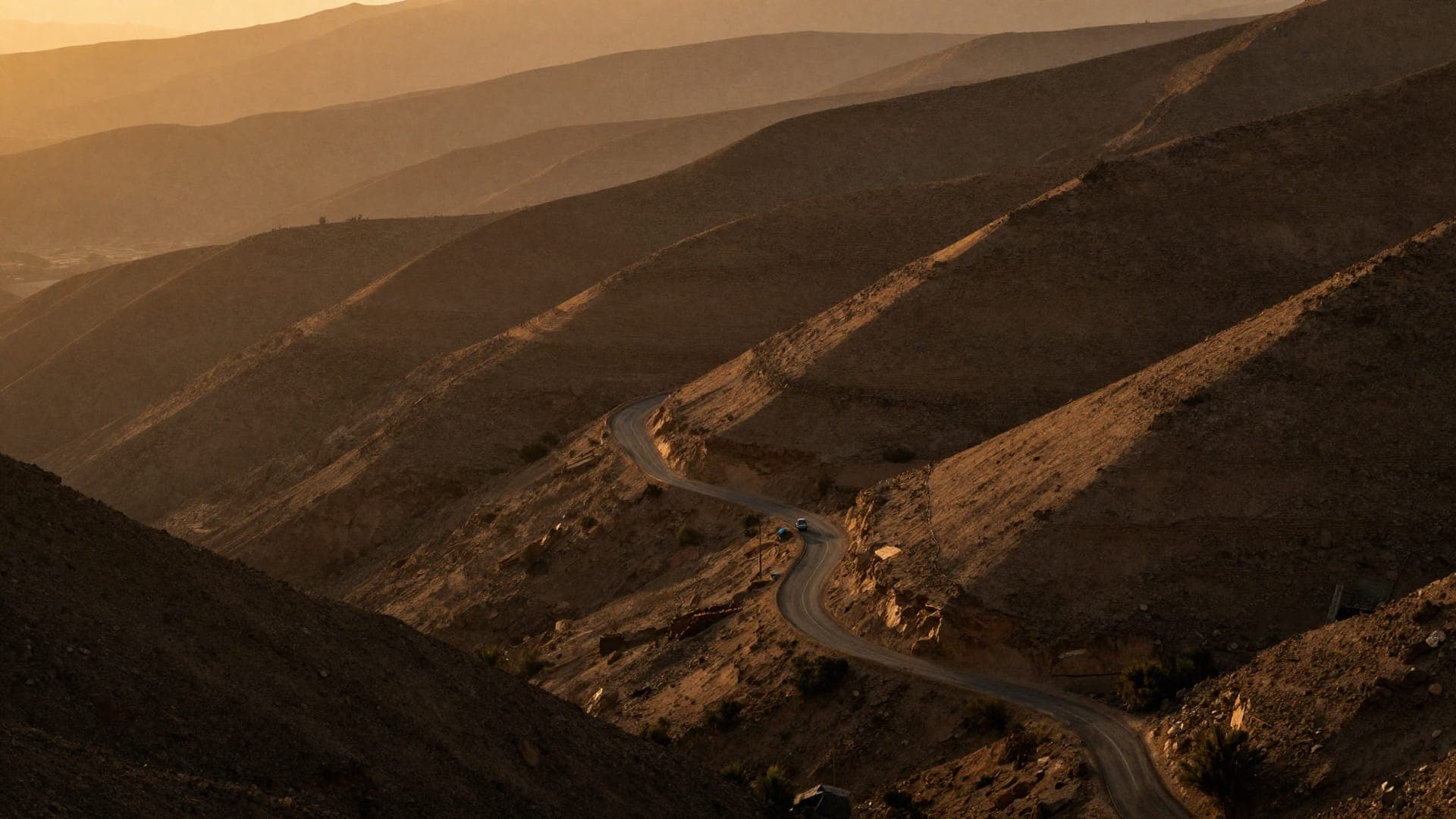 A single-track road folding back on itself down a flank of bare, sun-raked hills at sunset, ridge behind ridge stacking away into the haze.